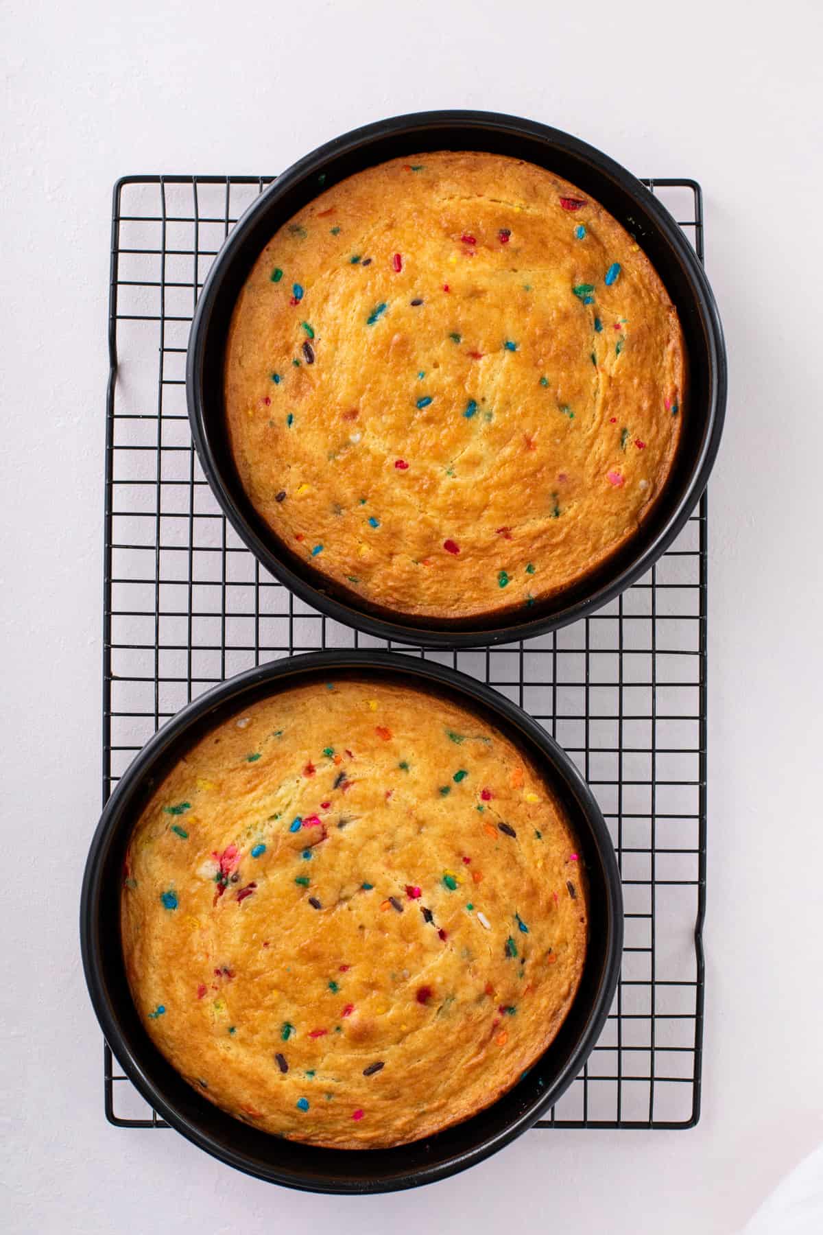 Two funfetti cakes in round cake pans cooling on a wire rack.