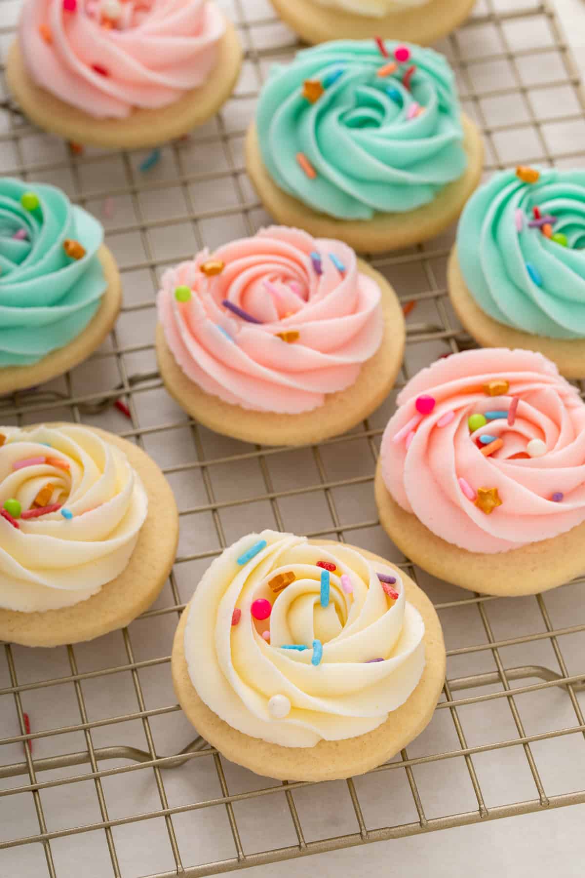 Sugar cookie frosting piped into rosettes in three different colors and topped with rainbow sprinkles on round sugar cookies that are resting on a wire rack.