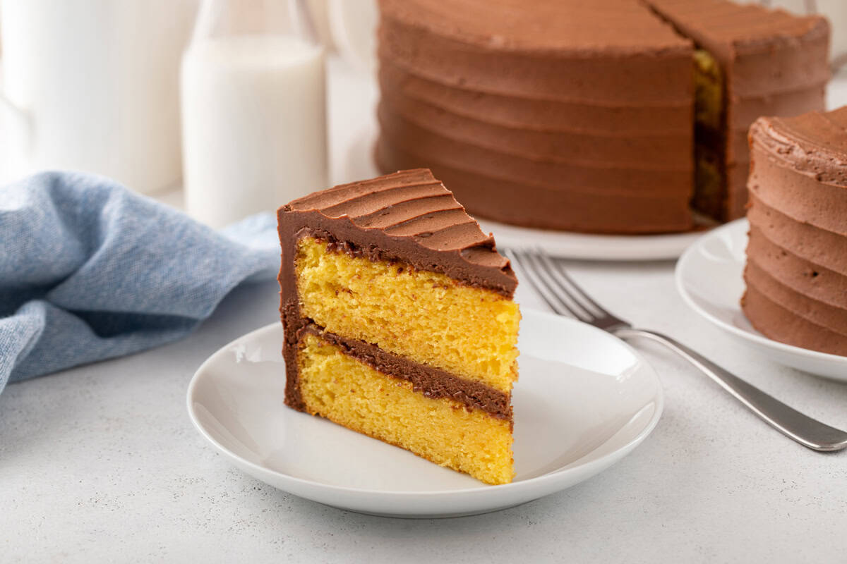 Slice of yellow layer cake with chocolate frosting on a white plate.