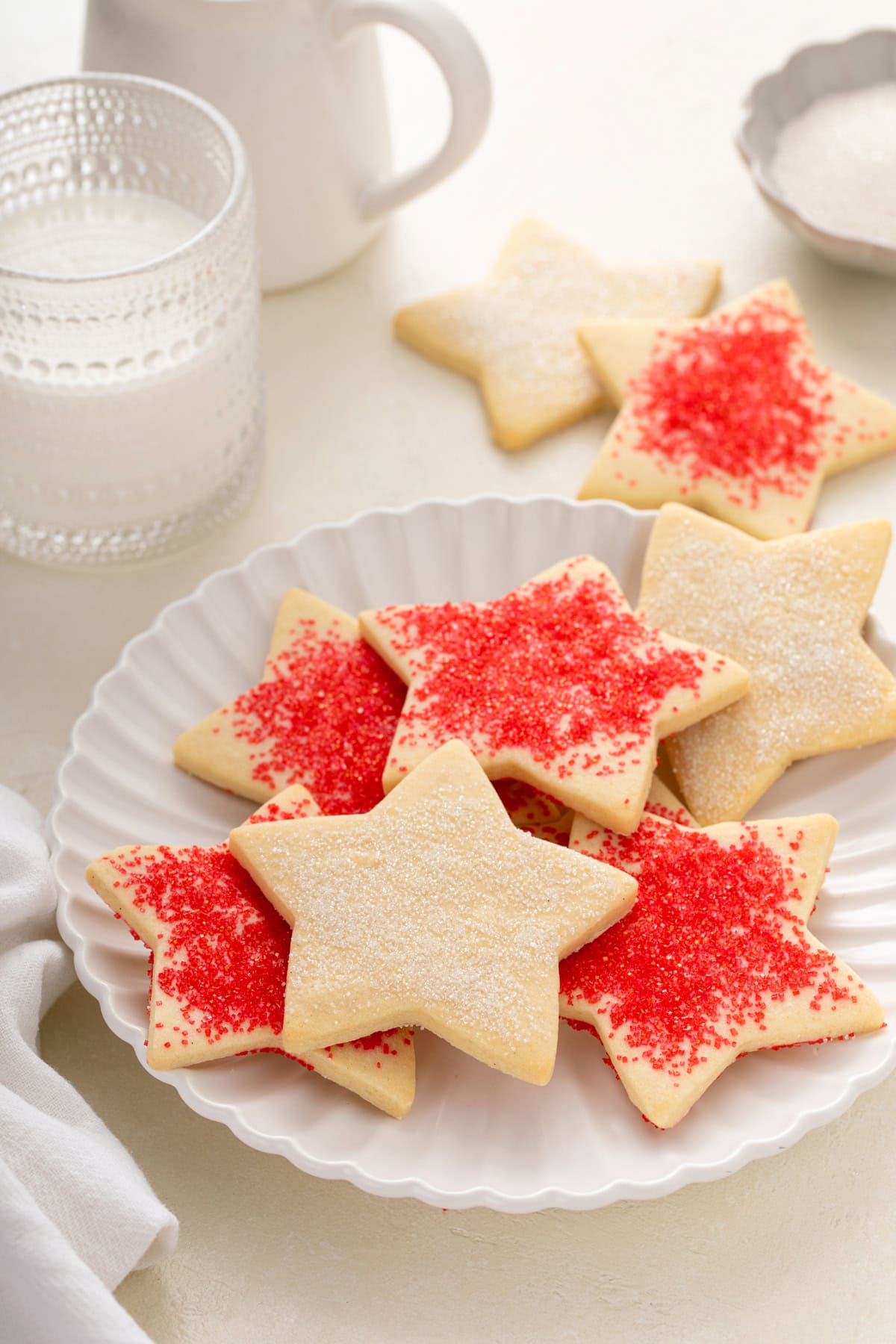 Star-shaped sugar cookies decorated with red and white sparkling sugar arranged on a white plate with a glass of milk in the background.