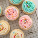 Sugar cookie frosting piped into rosettes in three different colors and topped with rainbow sprinkles on round sugar cookies.