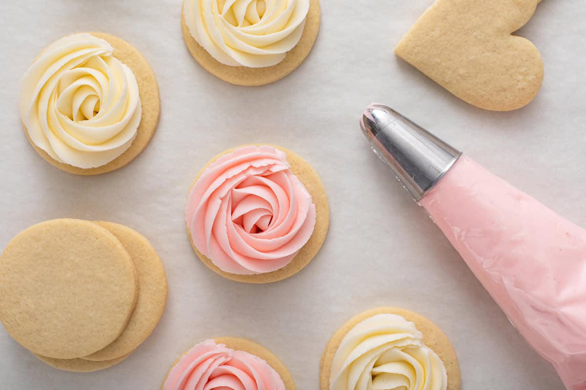 Overhead view of sugar cookie frosting piped in rosettes on round cookies, with a piping bag of pink frosting next to the cookies.