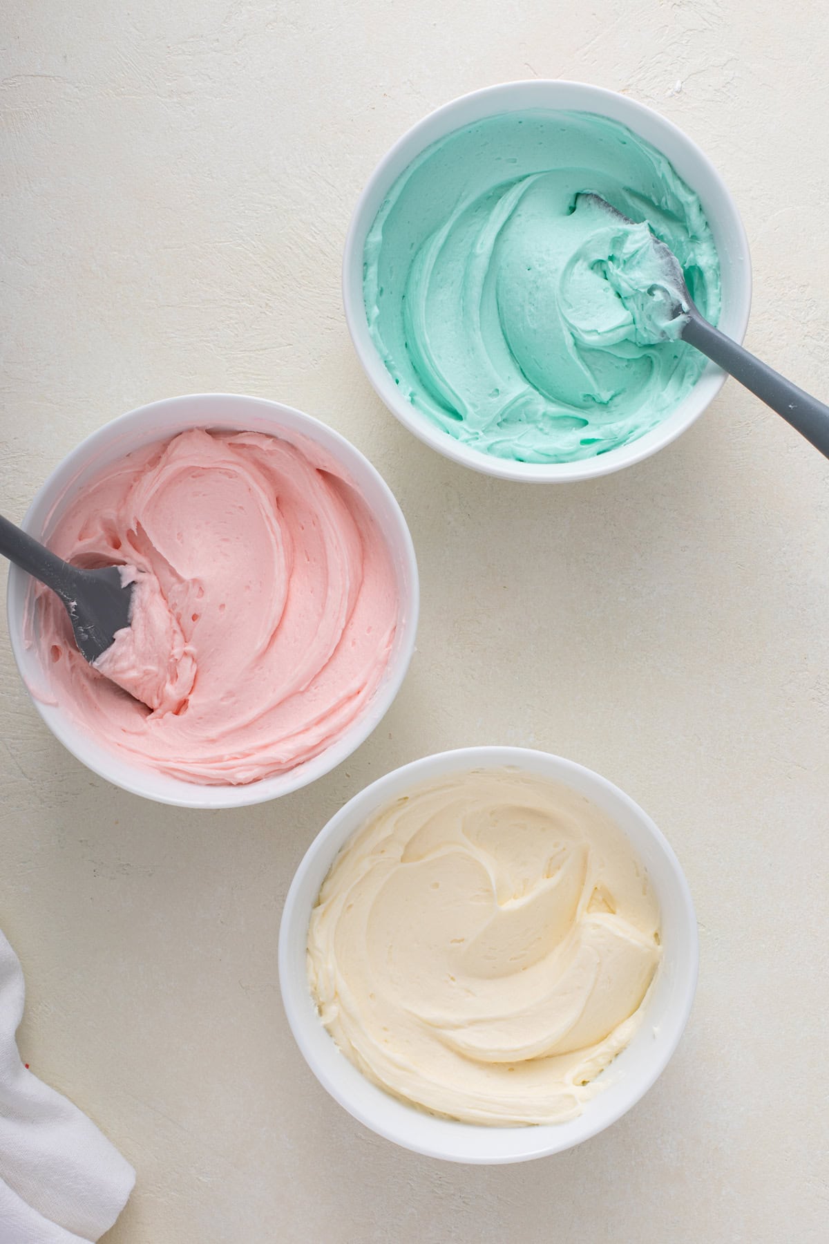 Three white bowls each filled with sugar cookie frosting. One bowl is left white and the others are being colored pink and mint green.