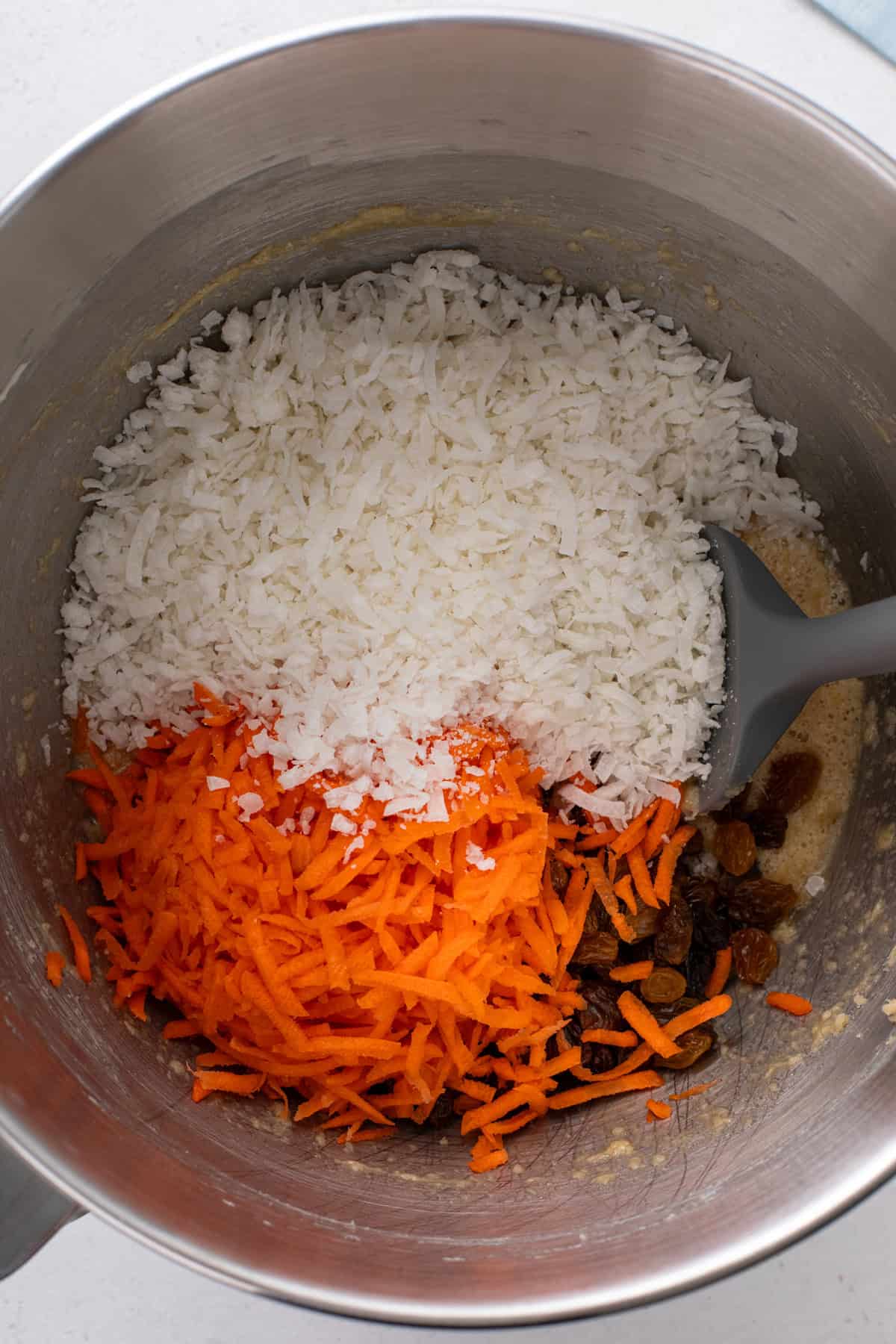 Shredded carrot, raisins, and coconut added to the bowl of a stand mixer.
