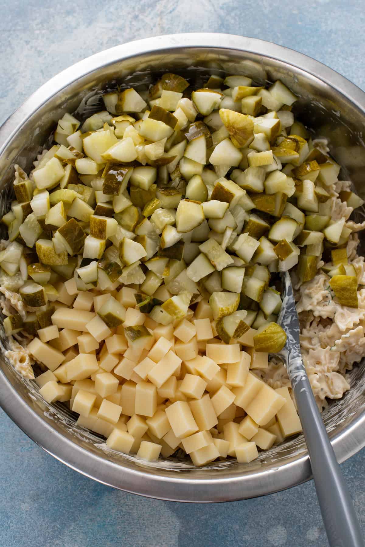 Chopped dill pickles and cubed cheese being added to a large mixing bowl.